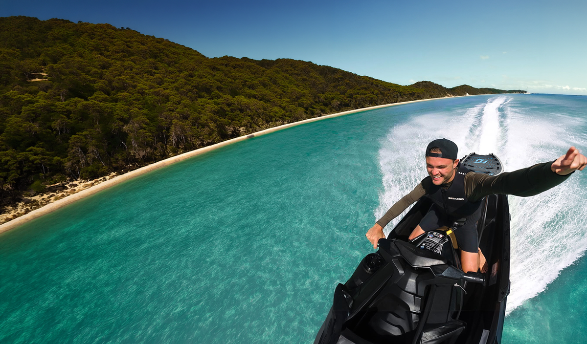 A man wearing a black wetsuit and cap rides a black jet ski on turquoise ocean water, with a sandy beach and dense green forest on the left and a blue sky above.